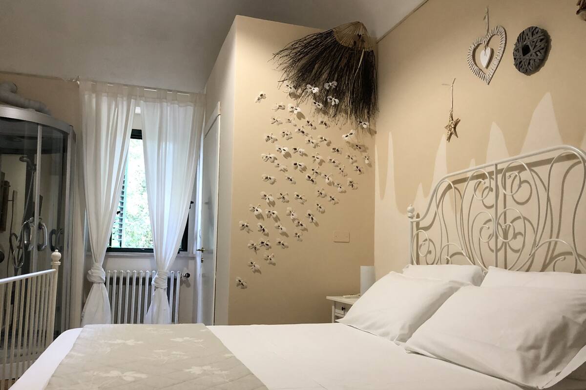 Un dormitorio con cama, ventana con cortinas, radiador y adornos de pared que incluyen un objeto con forma de corazón y una estrella.