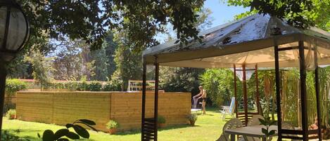A covered outdoor seating area with a table and chairs, surrounded by a wooden fence and greenery.