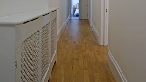 Um corredor com piso de madeira, paredes brancas e um radiador. Há quadros nas paredes e uma porta no final.