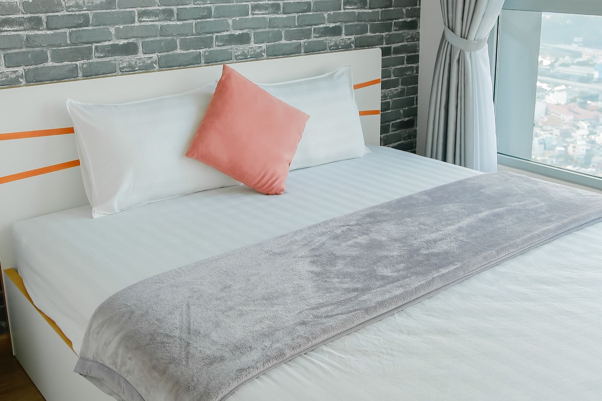 Una cama con ropa de cama blanca y un plaid gris, un cojín rosa y un cojín blanco con rayas anaranjadas. Encima de la cama, hay fotografías enmarcadas de monumentos famosos.