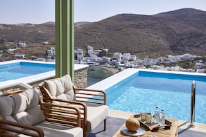 A poolside area with a view of a coastal village, a wooden lounge chair, and a tray with a bottle and glasses. - EmGard Suites Kythnos (Kithnos)