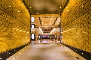 A corridor with a brick wall on one side and a reflective floor, leading to a seating area with tables and chairs.