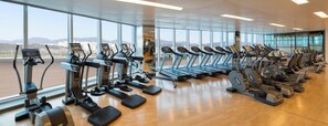 A gym with multiple treadmills and elliptical machines, large windows, and a wooden floor.