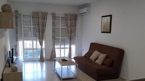 A living room with a brown sofa, a coffee table, and a TV on a stand. There is a window with curtains and an air conditioning unit on the wall. - Hospedería IslaSol (San Fernando)