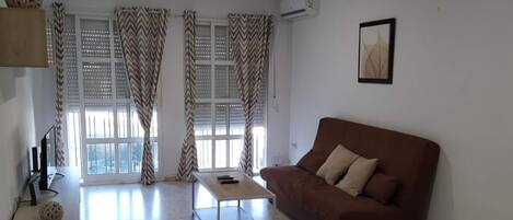 A living room with a brown sofa, a coffee table, and a TV on a stand. There is a window with curtains and an air conditioning unit on the wall.