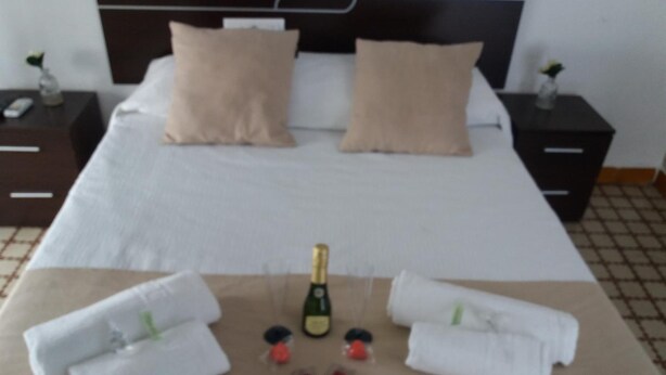 A neatly made bed with two pillows, a bottle of champagne, and two towels on a brown mat. - Hospedería IslaSol (San Fernando)