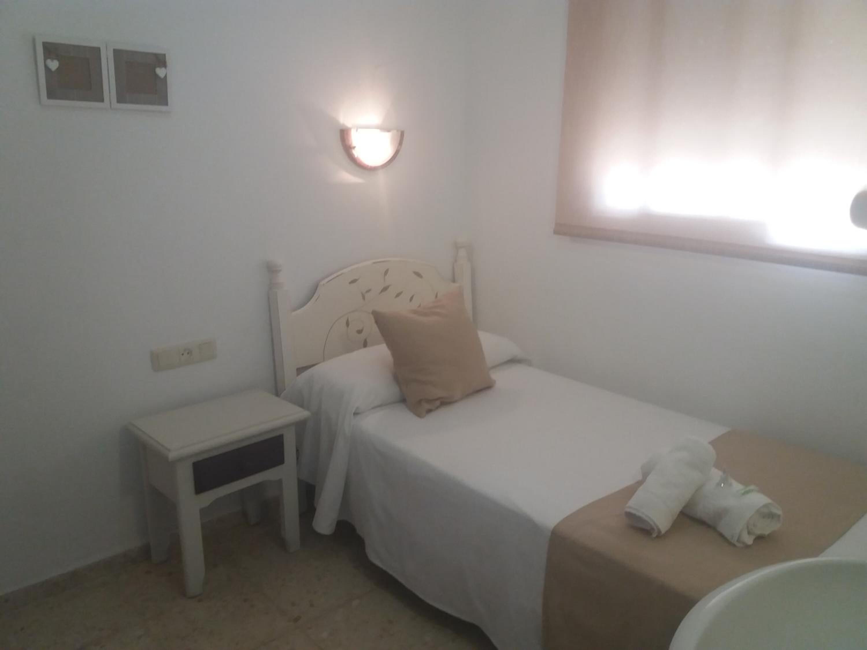 A small, neatly arranged bedroom with a single bed, a bedside table, a nightstand, and a wall-mounted light fixture.