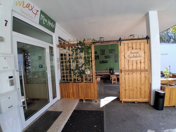 Entrada de um hostel com um portão de madeira identificado como "Annas Backpacktube", uma parede verde com um quadro de cardápio e uma área de estar com bancos.