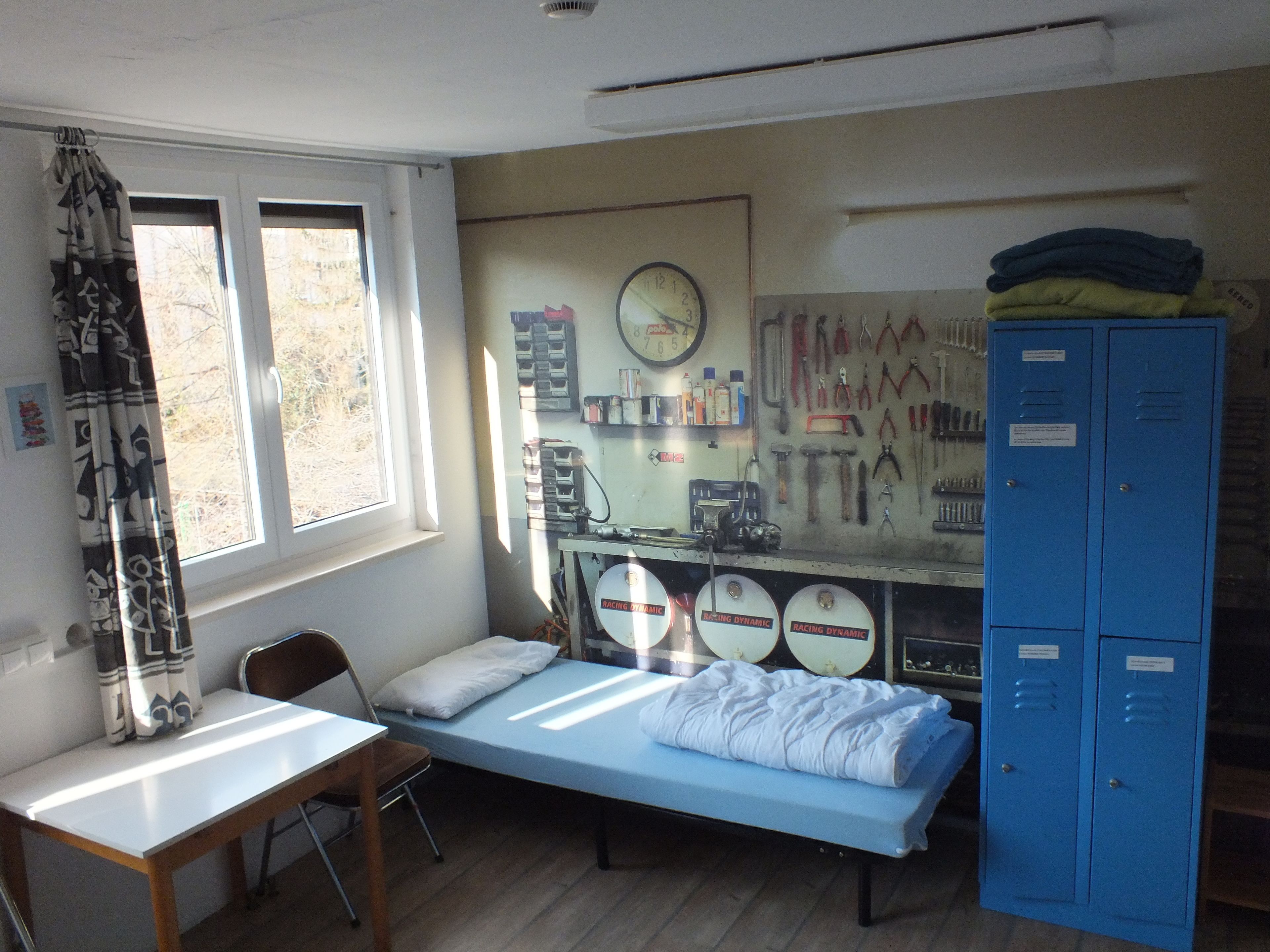 A dormitory room with a bed, a desk, a chair, a window, a wall clock, and a blue storage unit.