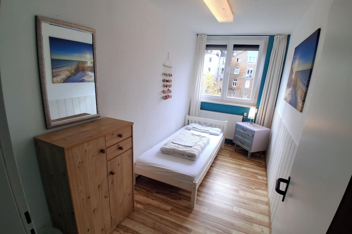 A bedroom with a wooden dresser, a bed, a bedside table, and a window with a view of buildings.