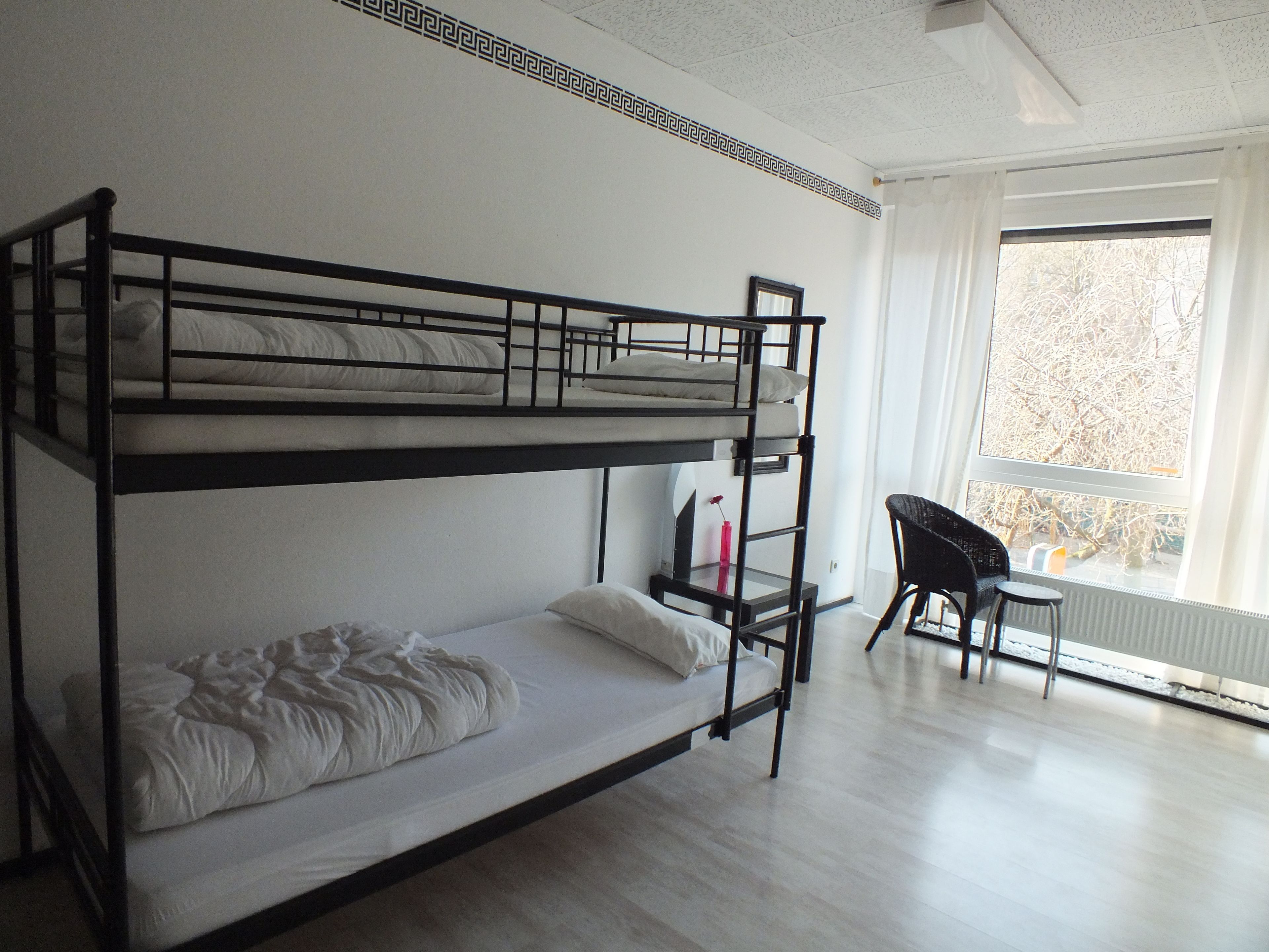 Een slaapkamer met een stapelbed, bureau en stoel, een radiator en een raam met uitzicht op bomen.