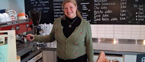 A person standing behind a counter with a coffee machine, a menu board, and a display of lollipops.