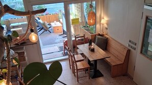 A cozy cafe with wooden furniture, large windows, and hanging plants.