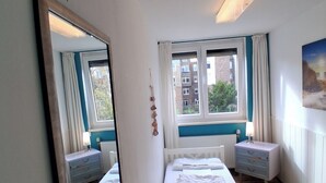 A bedroom with a large mirror, a wooden desk, a bed with white bedding, a window with curtains, and a radiator.