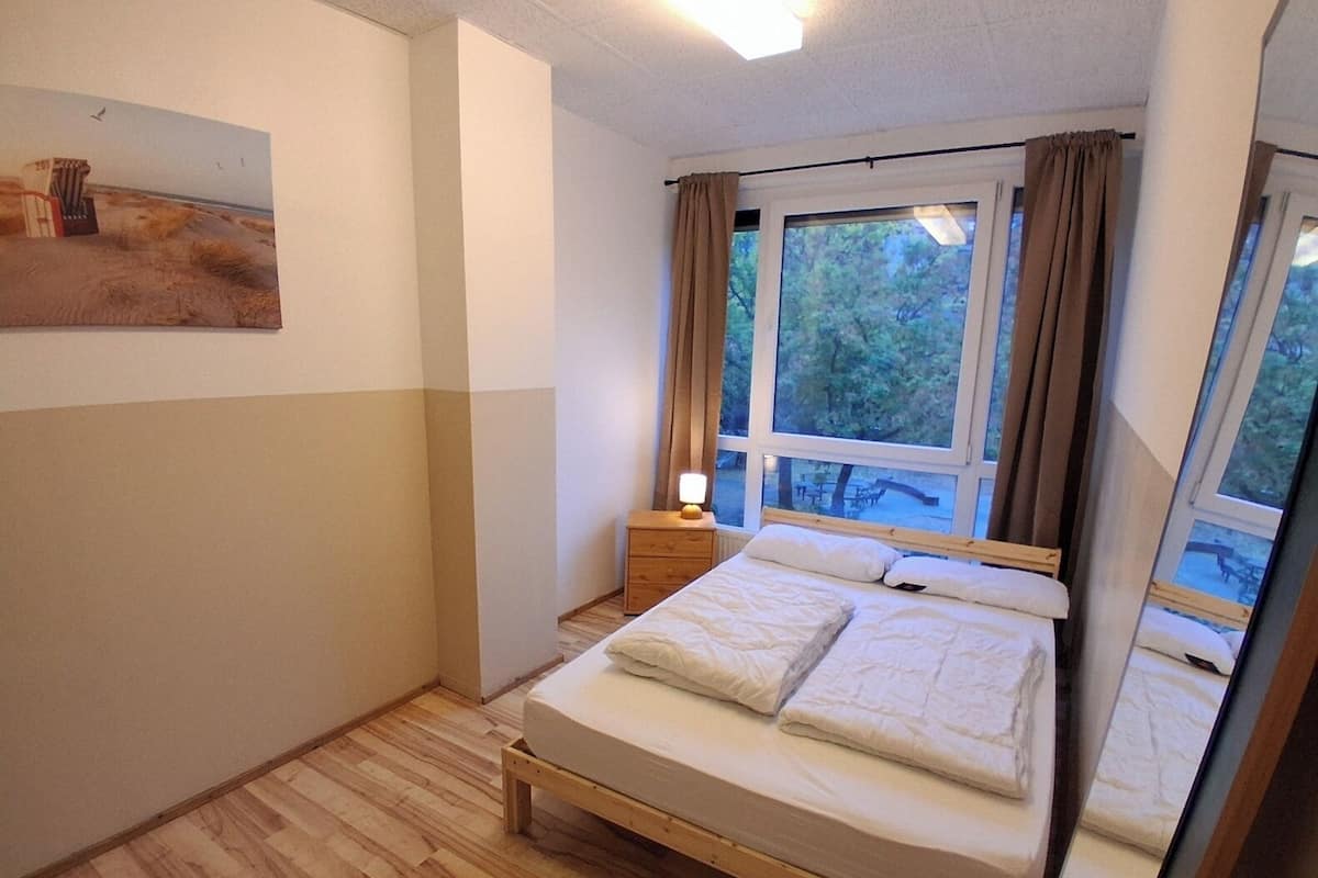 A bedroom with a wooden bed, a nightstand, a lamp, and a large window with curtains.