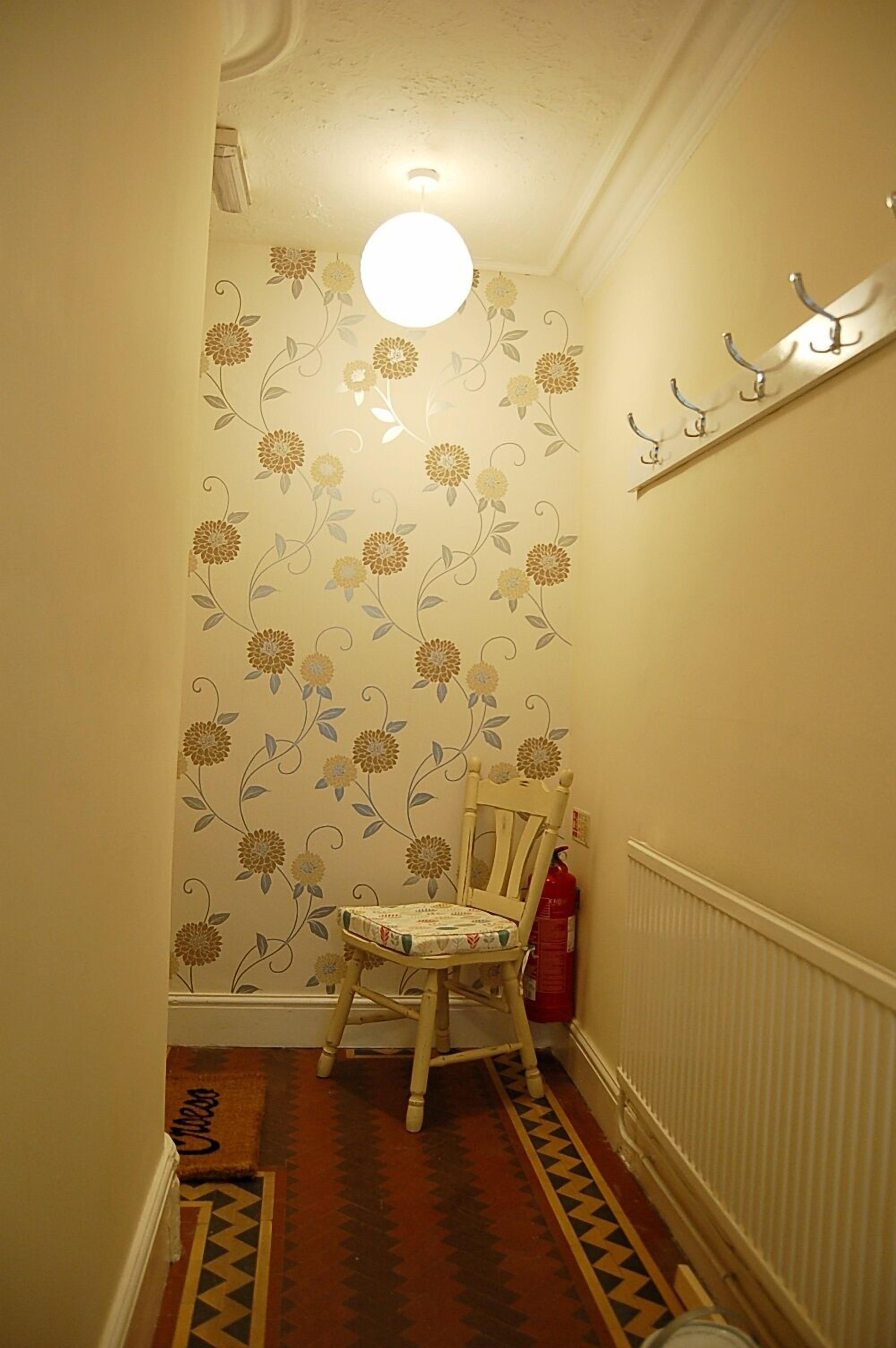 A narrow hallway with patterned wallpaper, a wooden chair, a radiator, and a small shelf with hooks.