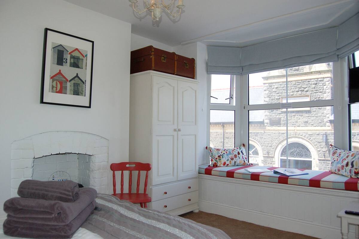 A bedroom with a bed, a red chair, a white wardrobe, a window seat with a quilt, and a framed picture on the wall.
