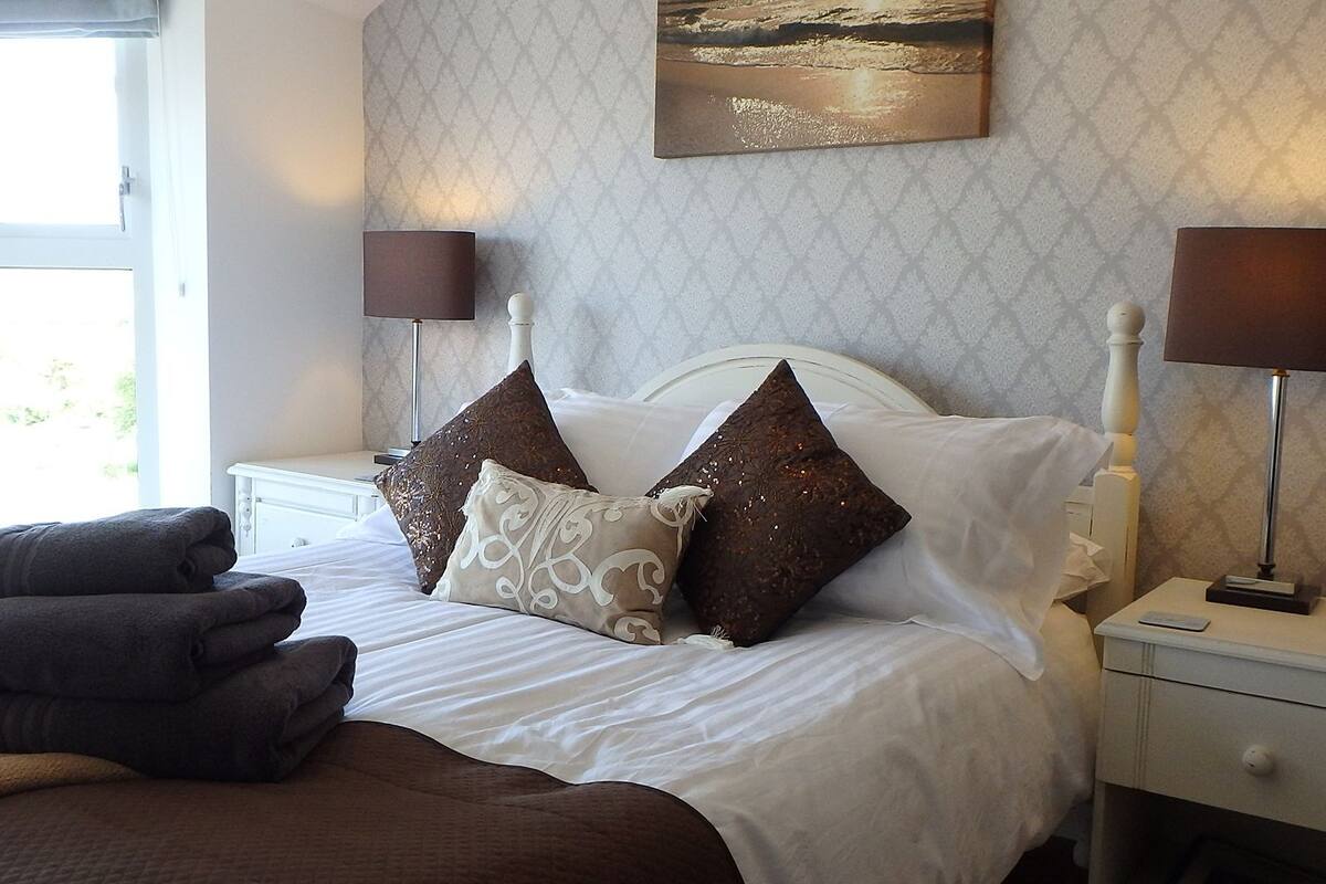 A neatly made bed with a white headboard, two bedside lamps, and a view of the sea through a window.