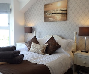 A neatly made bed with a white headboard, two bedside lamps, and a view of the sea through a window. - Saint Cadfans Lodge (Tywyn)