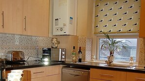 A kitchen with wooden cabinets, a dishwasher, a coffee maker, and a window with a plant. - Saint Cadfans Lodge (Tywyn)