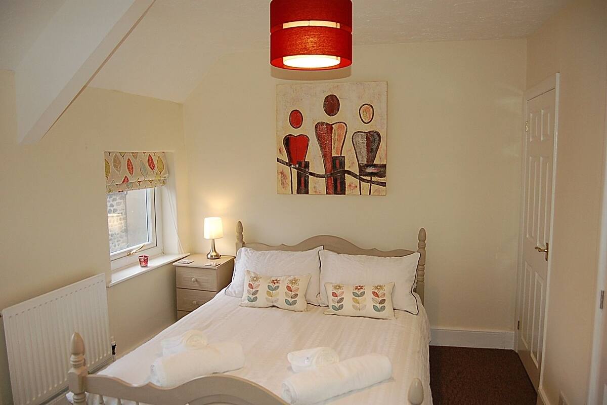 A bedroom with a bed, a nightstand, a window with curtains, a painting on the wall, and a door.