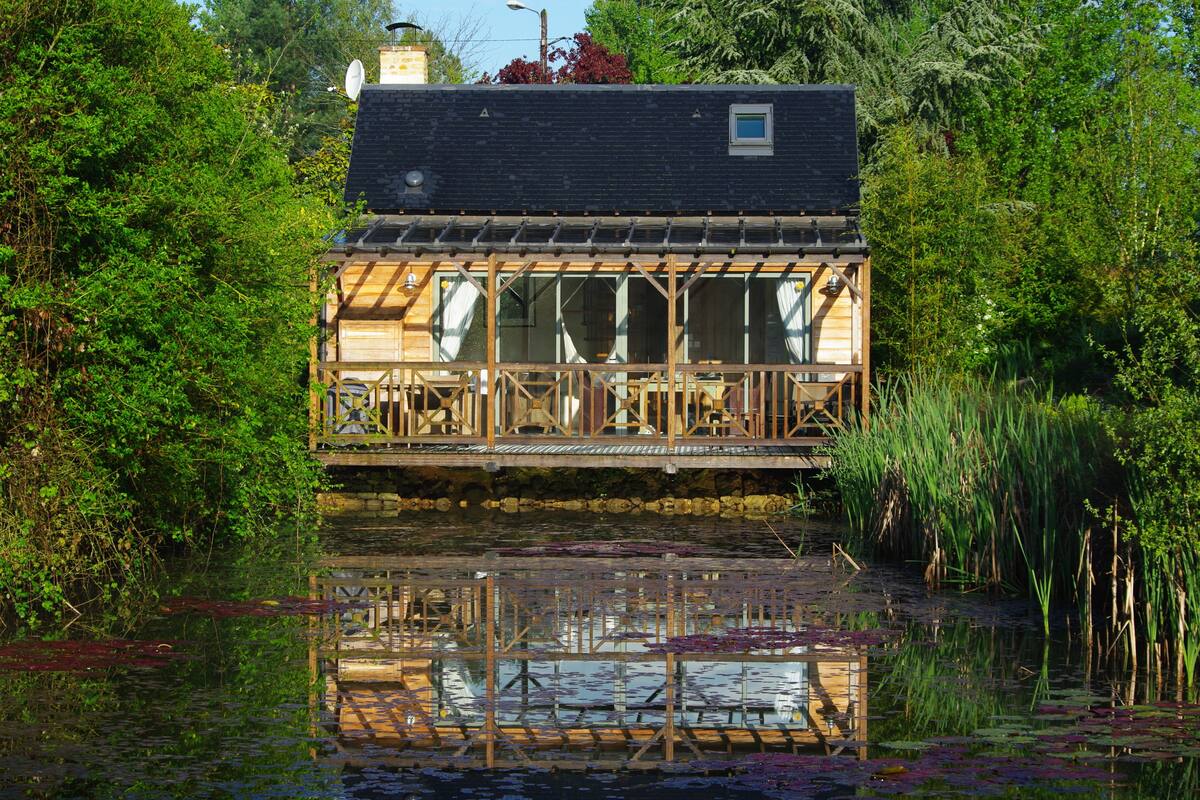 A wooden houseboat with a deck and windows, surrounded by water and greenery.