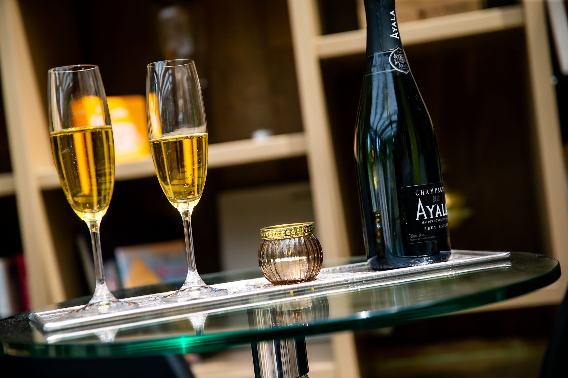 Two champagne glasses on a glass table with a bottle of Ayala Champagne and a small decorative jar.