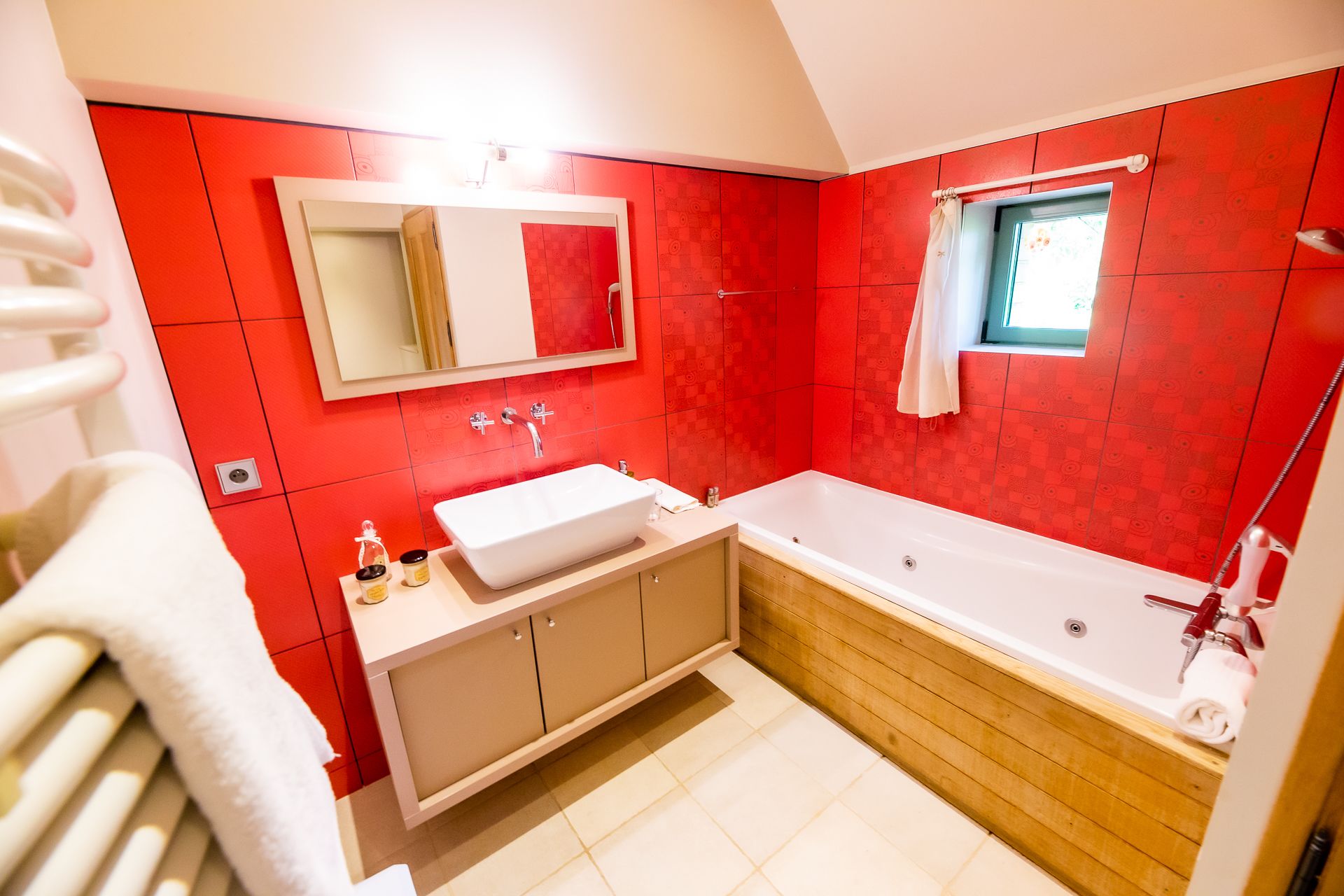 A modern bathroom with a red tiled wall, a white sink, a wooden vanity, and a bathtub.