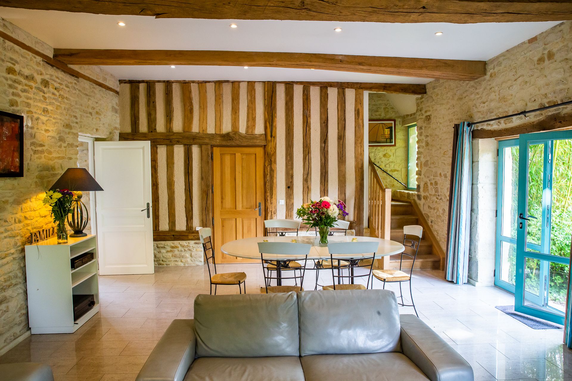 A cozy room with a stone wall, wooden beams, a dining table with chairs, a sofa, and a staircase leading to another level.