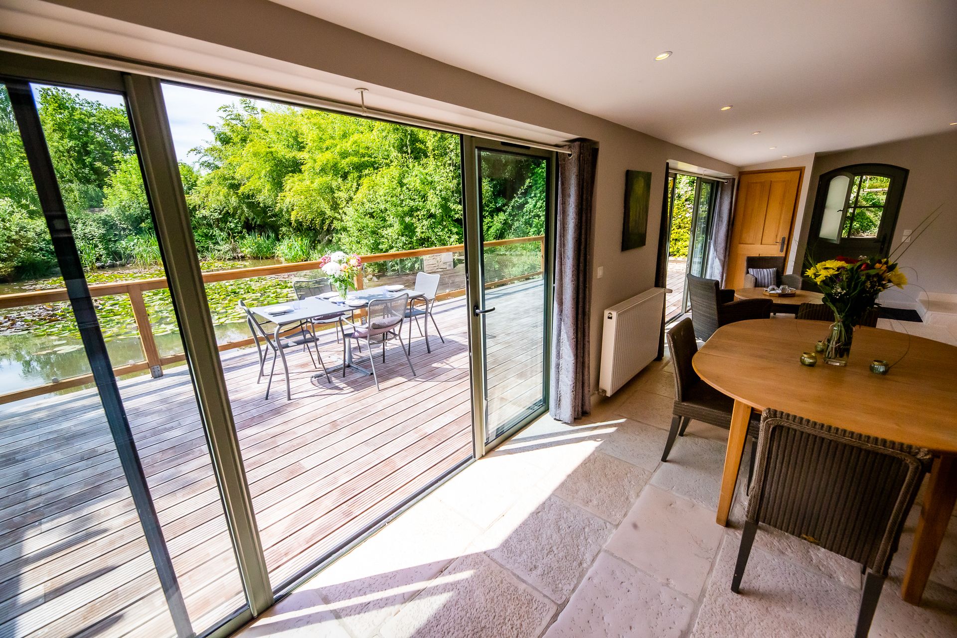A spacious room with a large sliding glass door leading to an outdoor deck with a wooden table and chairs, a view of greenery, and a small kitchen area with a round table and a vase of flowers.
