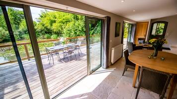 A spacious room with a large sliding glass door leading to an outdoor deck with a wooden table and chairs, a view of greenery, and a small kitchen area with a round table and a vase of flowers.