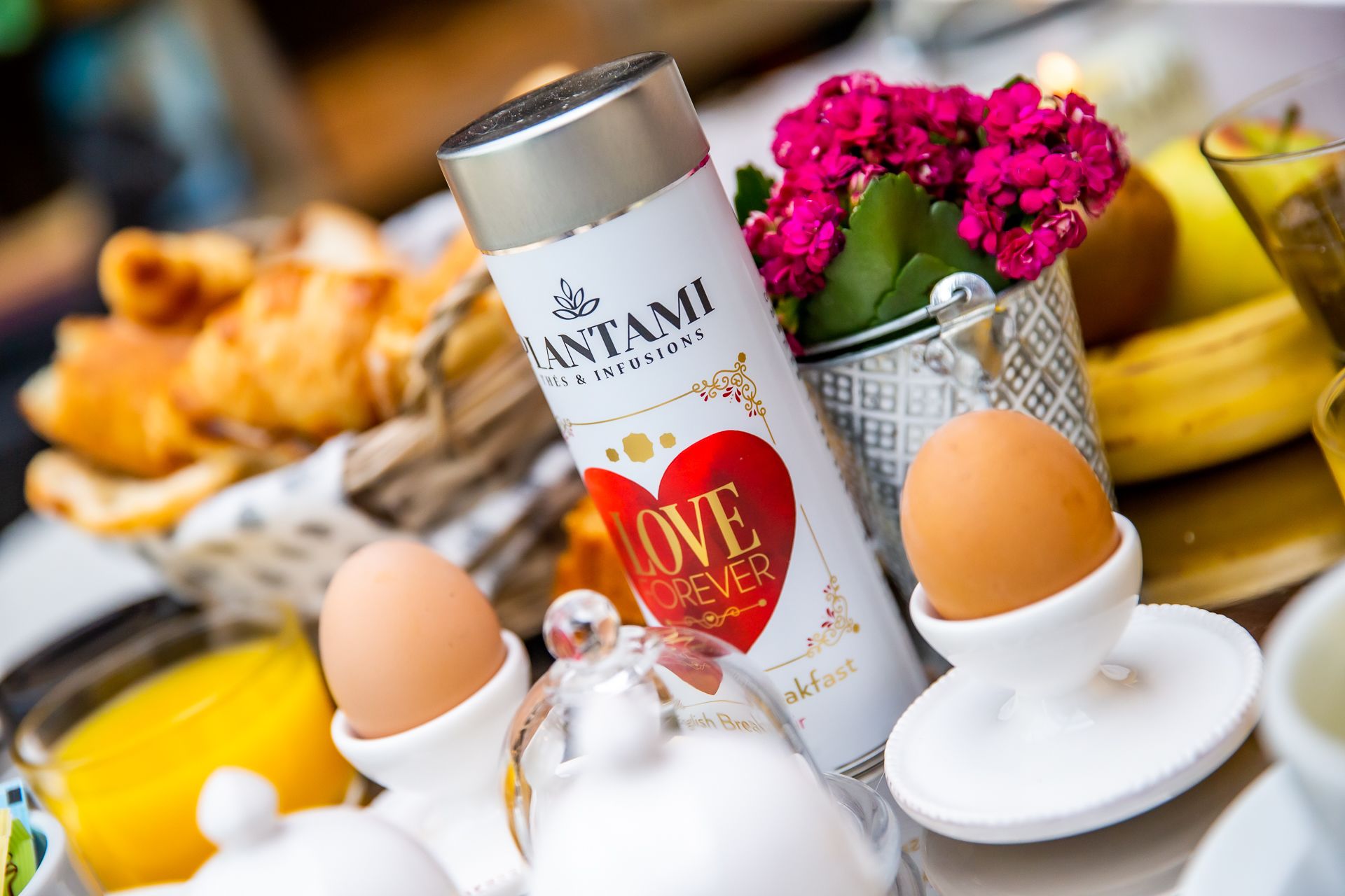 A breakfast setting with a can of plantami infusions, eggs, a glass of orange juice, and a basket of pastries.