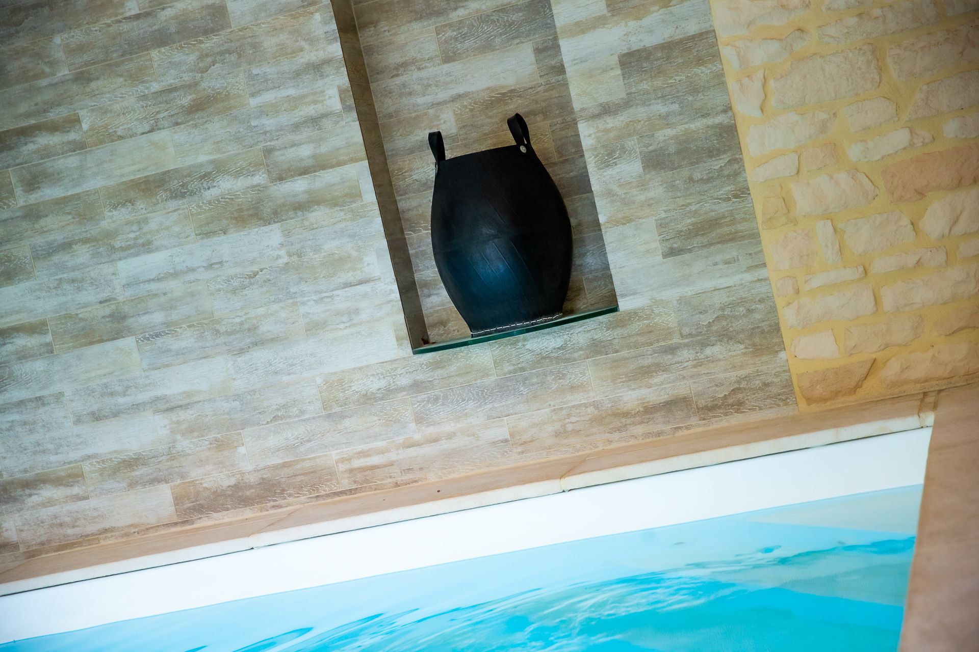 A black vase on a glass shelf against a textured wall.