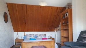 A bedroom with a wooden ceiling, a bed with patterned pillows, a bedside table with lamps, a colorful ottoman, a wooden wardrobe, and a sofa. - Chambres d'hôtes Maison Entre Terre et Mer (Primelin)