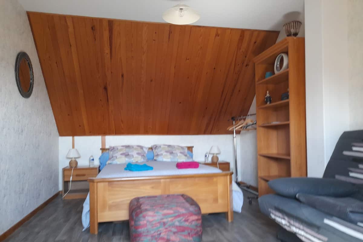 Un dormitorio con techo de madera, una cama con almohadones estampados, una mesita de noche con lámparas, un otomano colorido, un armario de madera y un sofá.