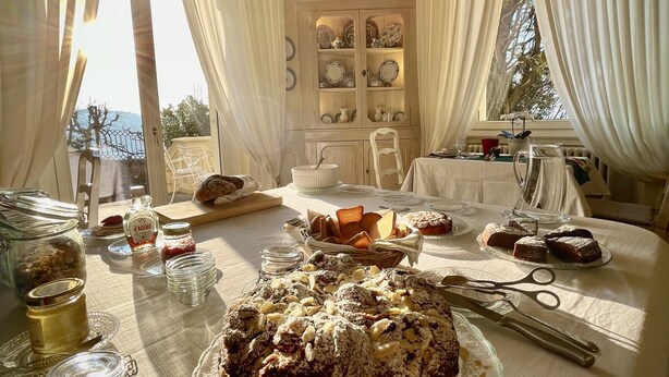 A dining area with a large cake on a glass stand, a table set with plates, glasses, and cutlery, and a view of the outdoors through large windows. - Villa La Moresca Relais de charme B&B adults only (Montecatini Terme)
