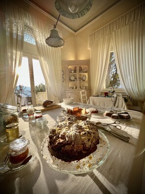A dining area with a large cake on a glass stand, a table set with plates, glasses, and cutlery, and a view of the outdoors through large windows. - Villa La Moresca Relais de charme B&B adults only (Montecatini Terme)