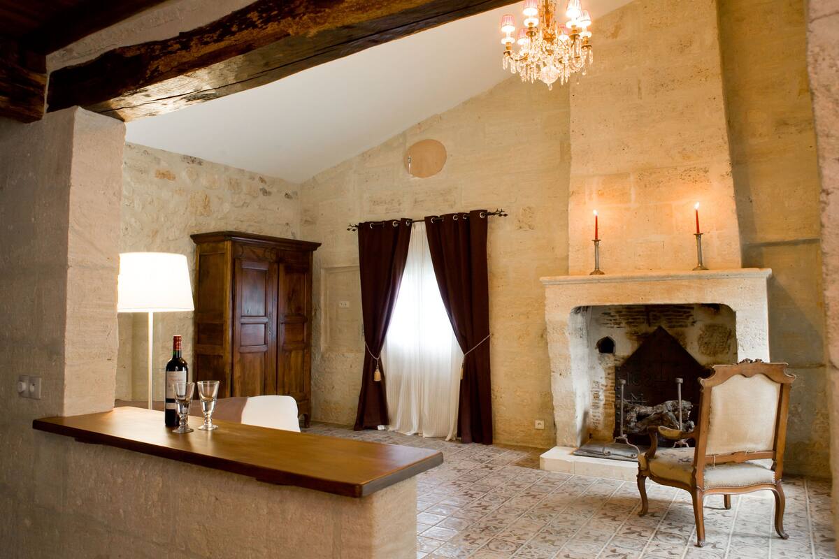 A rustic room with a fireplace, a wooden chair, a table with a bottle and glasses, and a chandelier.