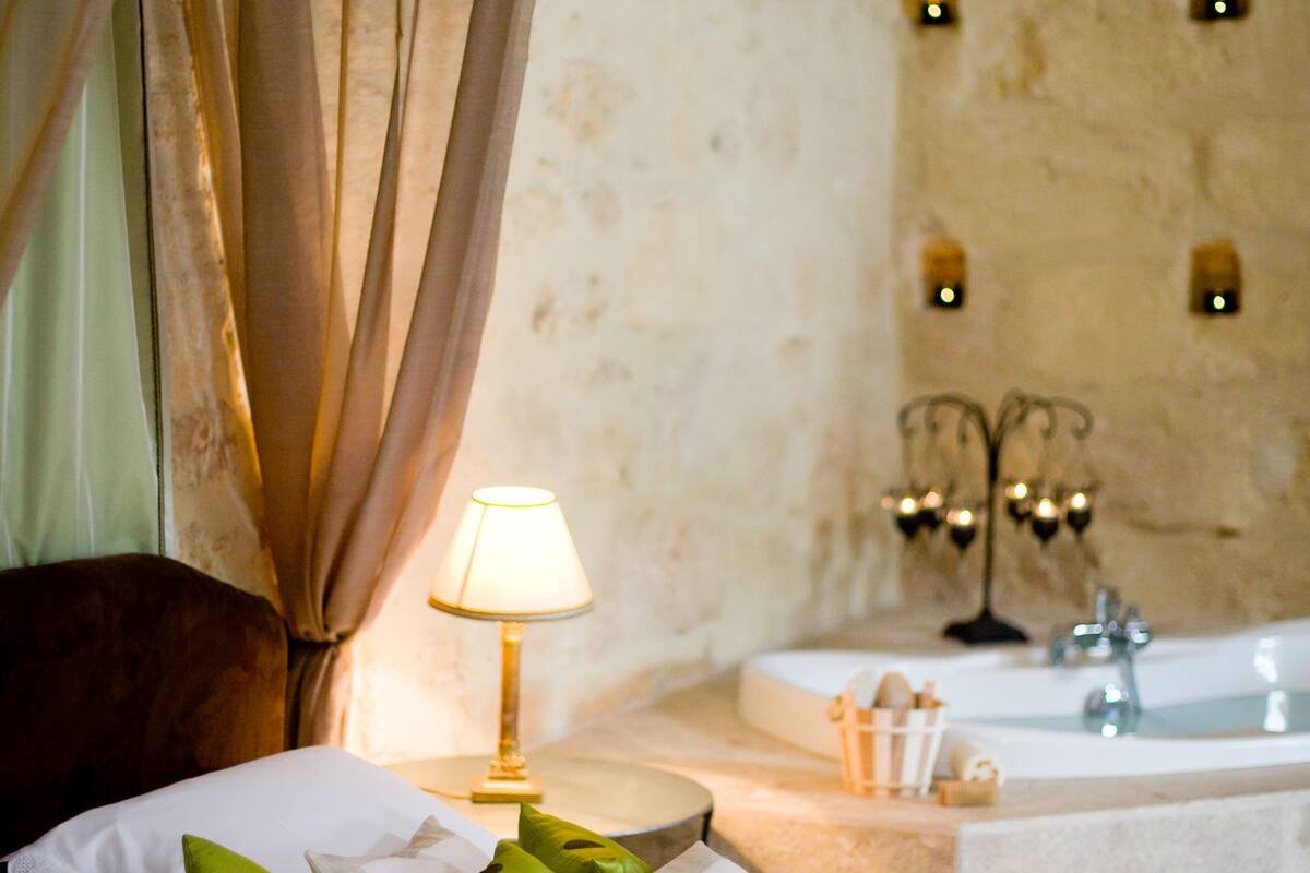 A hotel room with a bed, green pillows, a bedside lamp, a bathtub, and a stone wall with wall sconces.