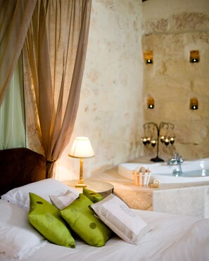 A hotel room with a bed, green pillows, a bedside lamp, a bathtub, and a stone wall with wall sconces.