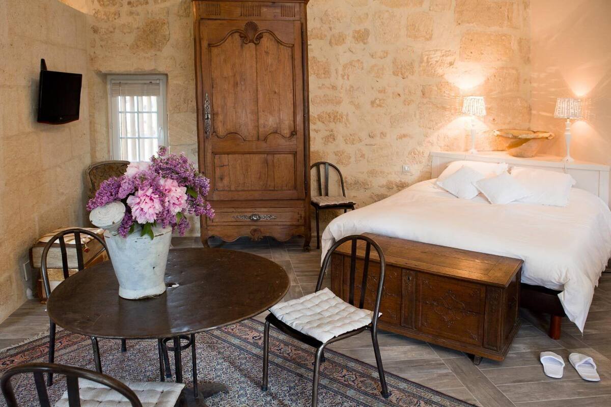 A bedroom with a bed, bedside lamps, a wooden wardrobe, a dining table with chairs, and a vase of flowers.