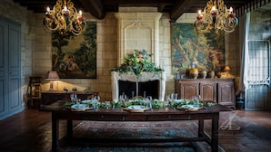 A dining room with a large wooden table set for a meal, flanked by two ornate chandeliers, a fireplace with a floral arrangement, and a stone wall with a mural. - Château Latour Segur Suites & Spa (Lussac)