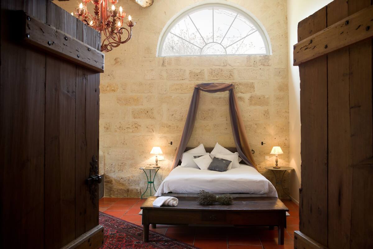 A bedroom with a four-poster bed, two bedside tables with lamps, a red patterned rug, and a large arched window.