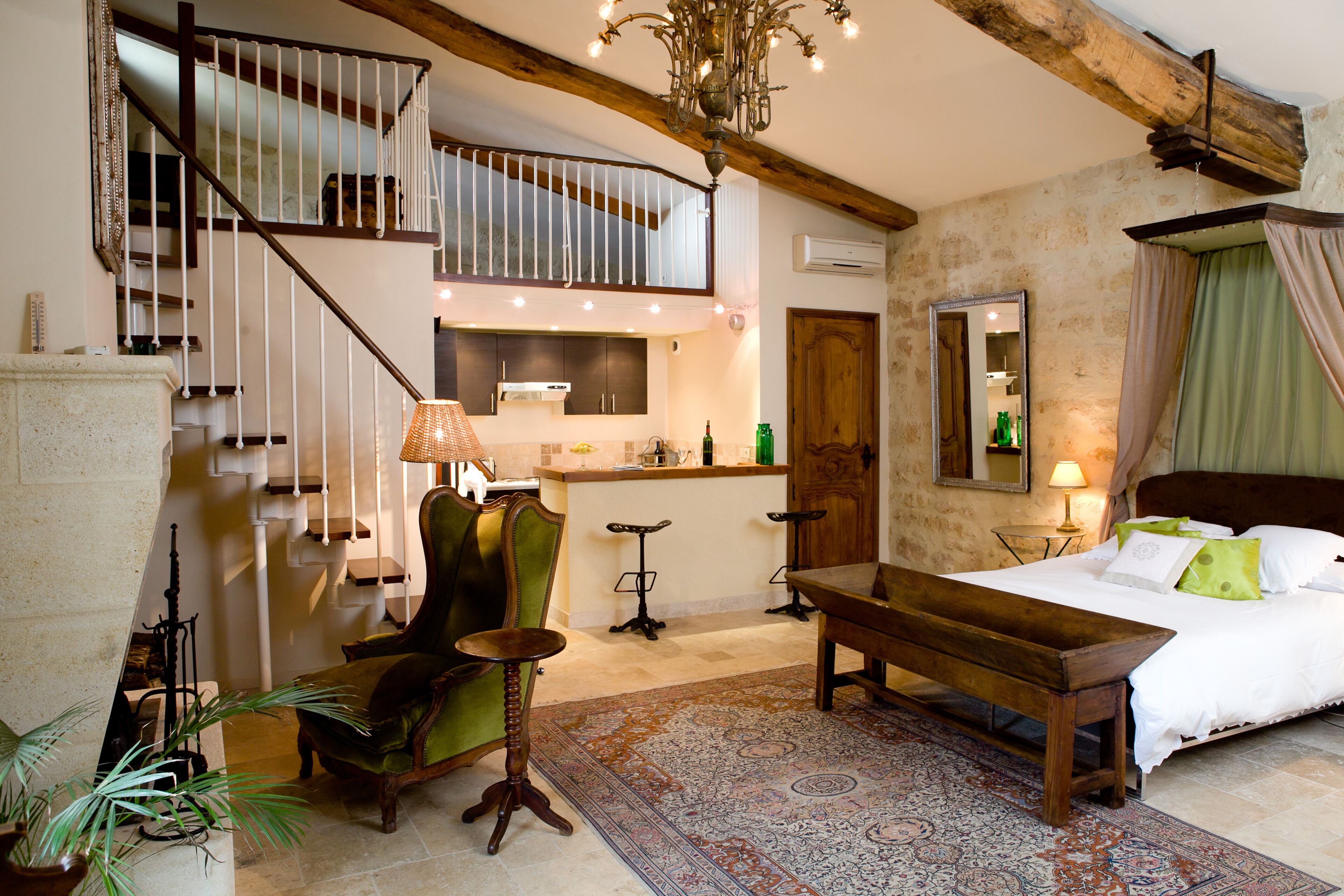 A bedroom with a large bed, a wooden bench, a green armchair, a dining table, a chandelier, a staircase, and a kitchen area.
