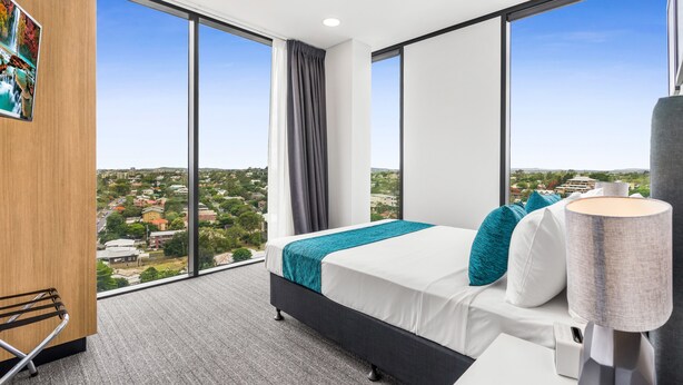 A modern hotel room with a large bed, a view of the city, and a cityscape through large windows. - Essence Suites Taringa (Taringa)