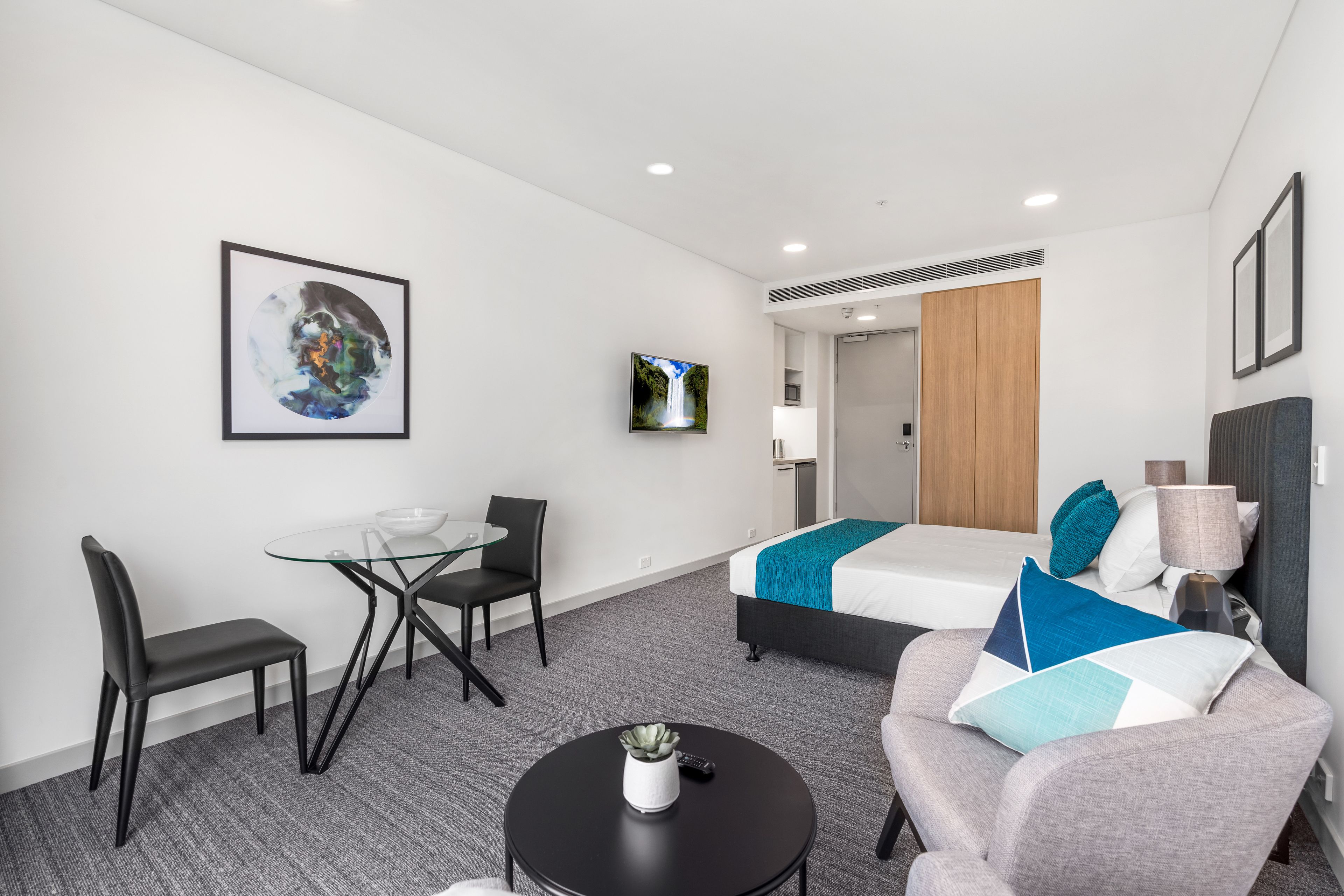 A modern hotel room with a glass dining table, two chairs, a sofa, a bed with blue and white bedding, a TV on the wall, and a small table with a lamp and remote control.