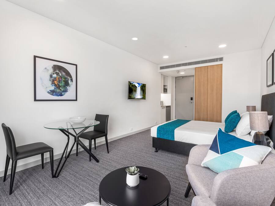 A modern hotel room with a glass dining table, two chairs, a sofa, a bed with blue and white bedding, a TV on the wall, and a small table with a lamp and remote control.