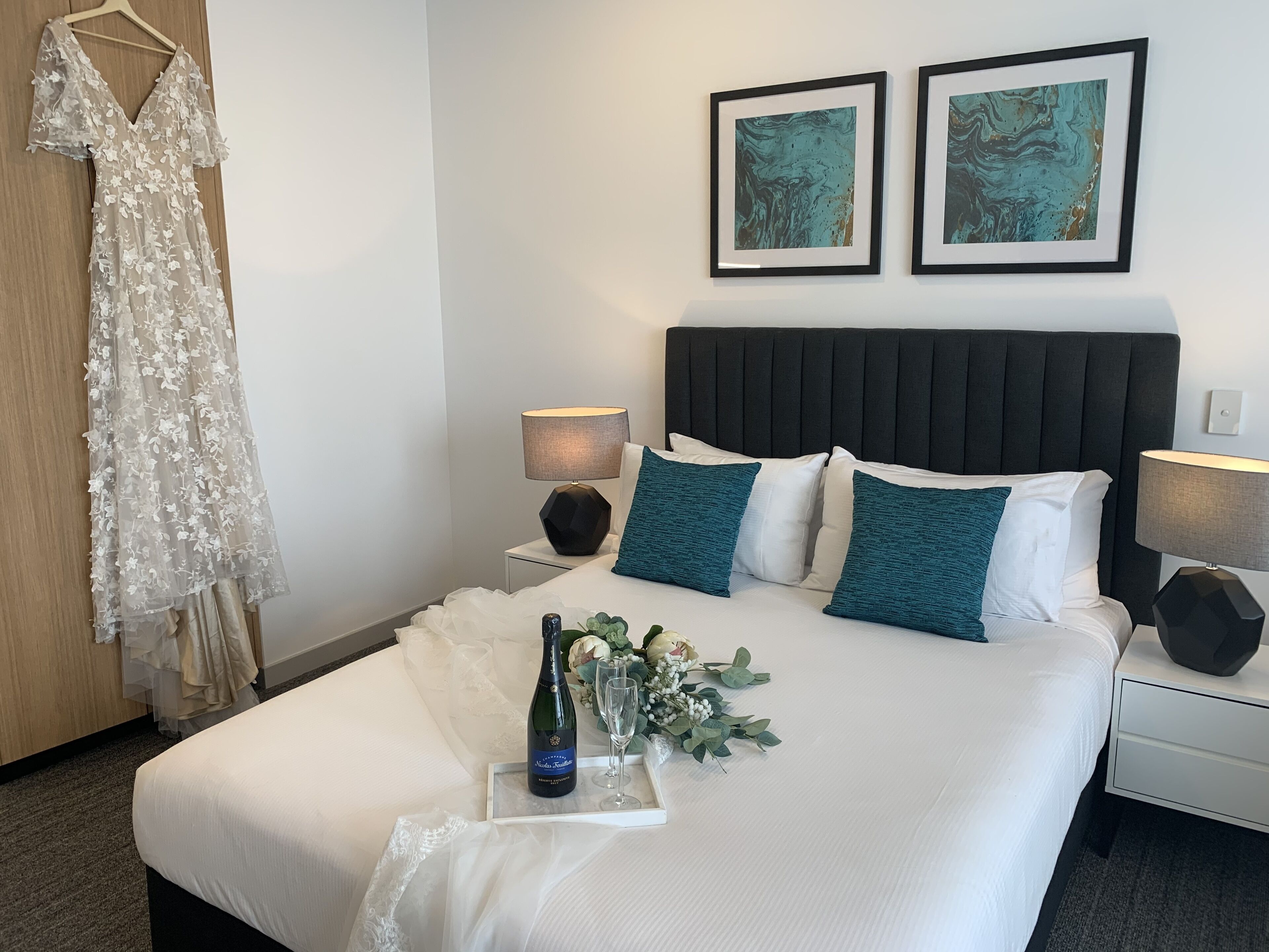 A hotel room with a bed, two framed abstract paintings, a nightstand with a lamp, a bottle of champagne, and a bouquet of flowers.
