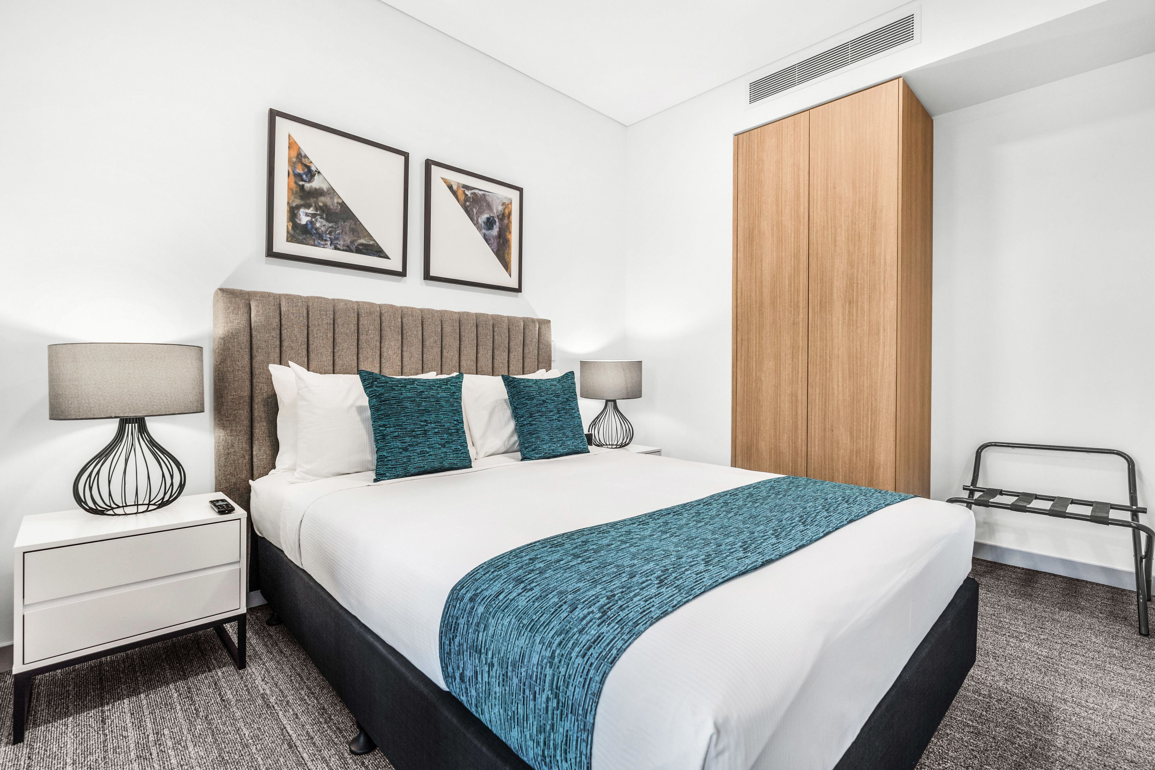 A modern hotel room with a large bed, two bedside tables with lamps, a wooden wardrobe, and two framed pictures on the wall.