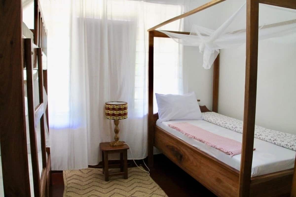 Una cama con dosel de madera con ropa blanca, una mesita de noche con lámpara y un ventanal con cortinas translúcidas.